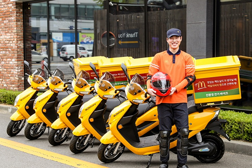 노란색 맥딜리버리 전기 배달 바이크 앞에 직원이 서 있는 모습으로, 친환경 배달 시스템을 상징함.